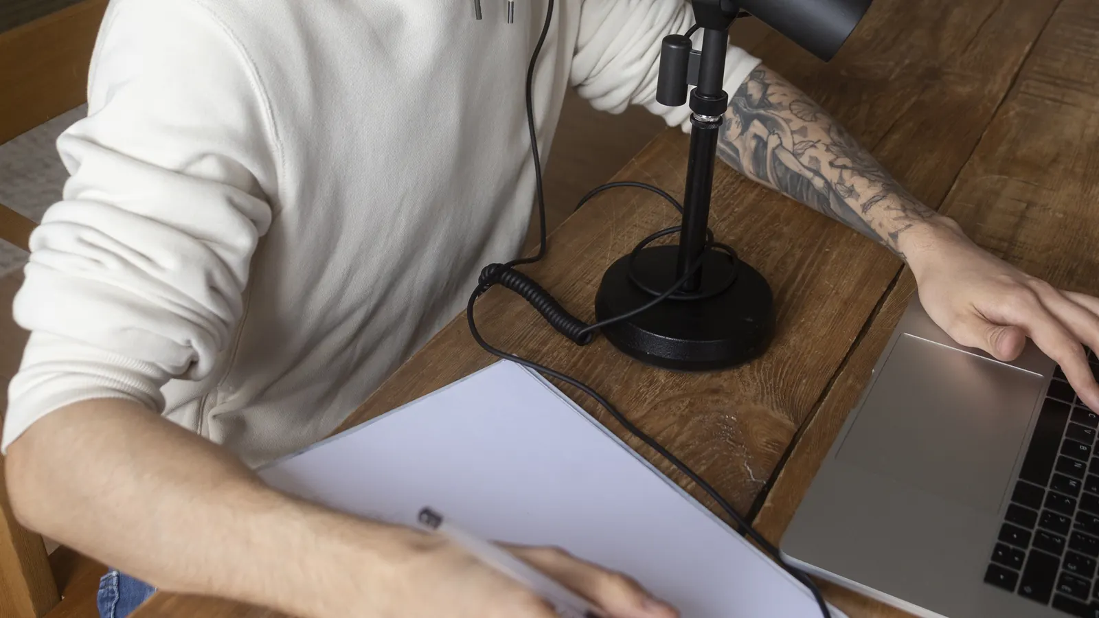 Seen from above, a man writes notes on a clipboard beside a desk microphone while his other hand rests on a laptop.
