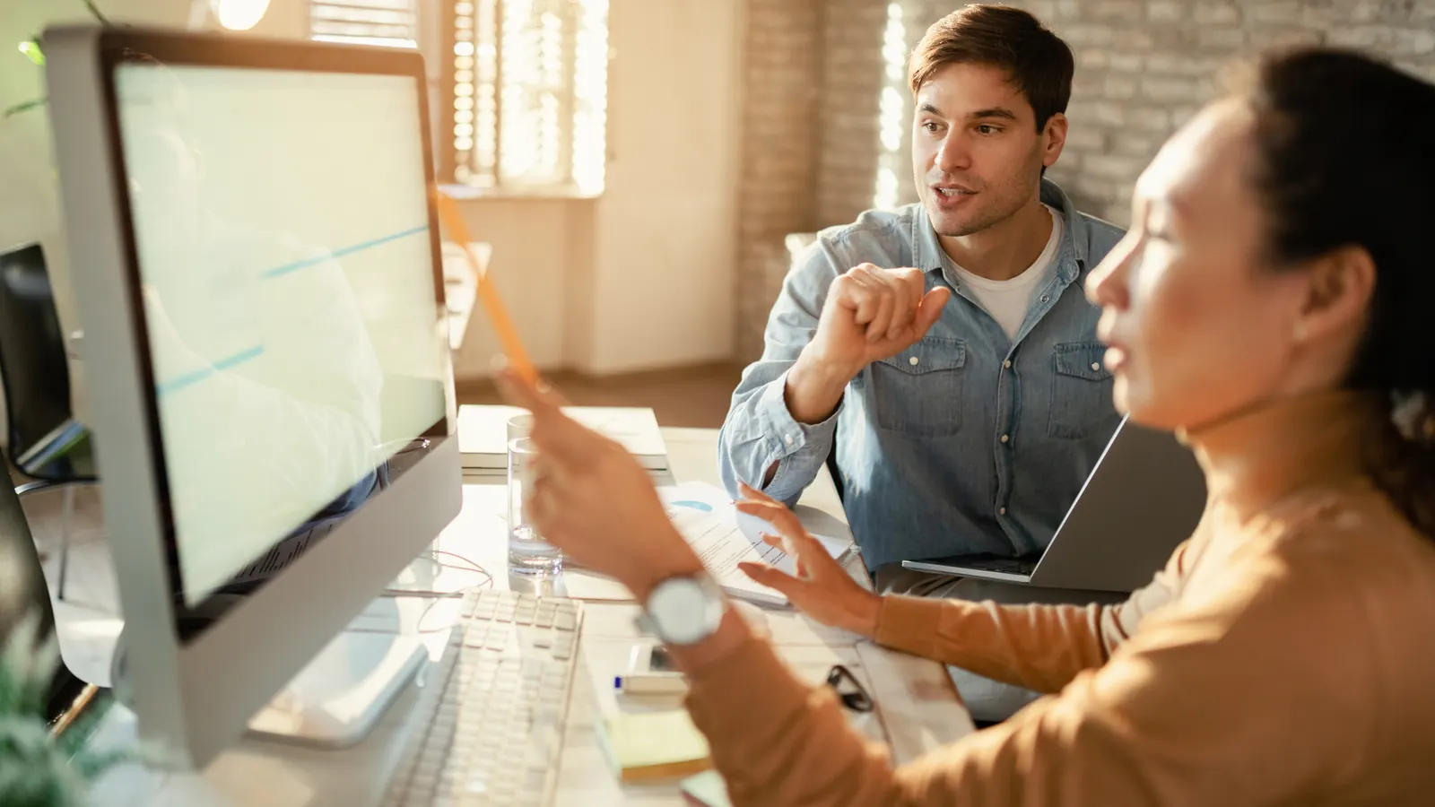 A woman points a pencil at a chart on a desktop monitor while a colleague in a denim shirt explains something across the desk.