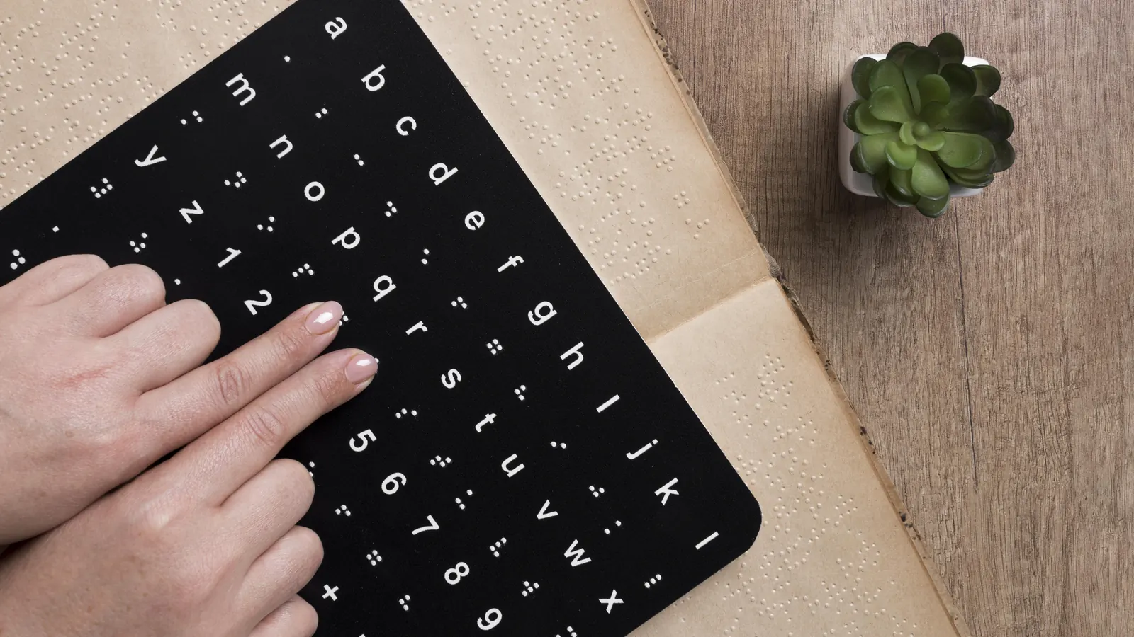 Two fingers touch the raised dots on a black braille alphabet board resting on an open braille book.