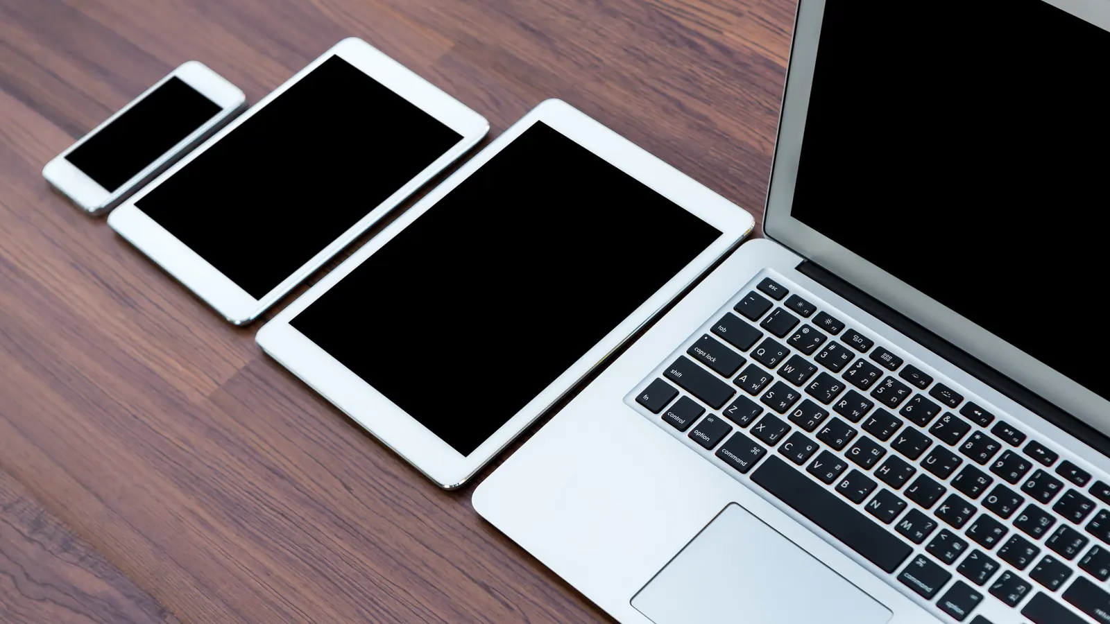 A smartphone, two tablets and a laptop lined up on a wooden table, all with dark screens.