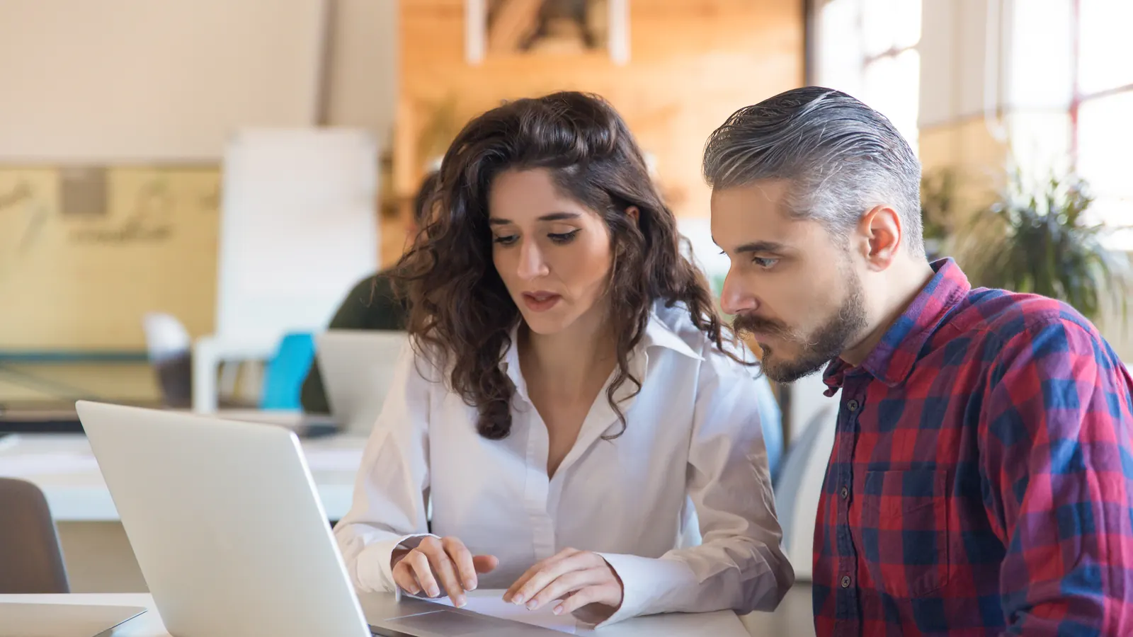 Two colleagues sit at a desk looking closely at a laptop screen, one typing while the other watches.